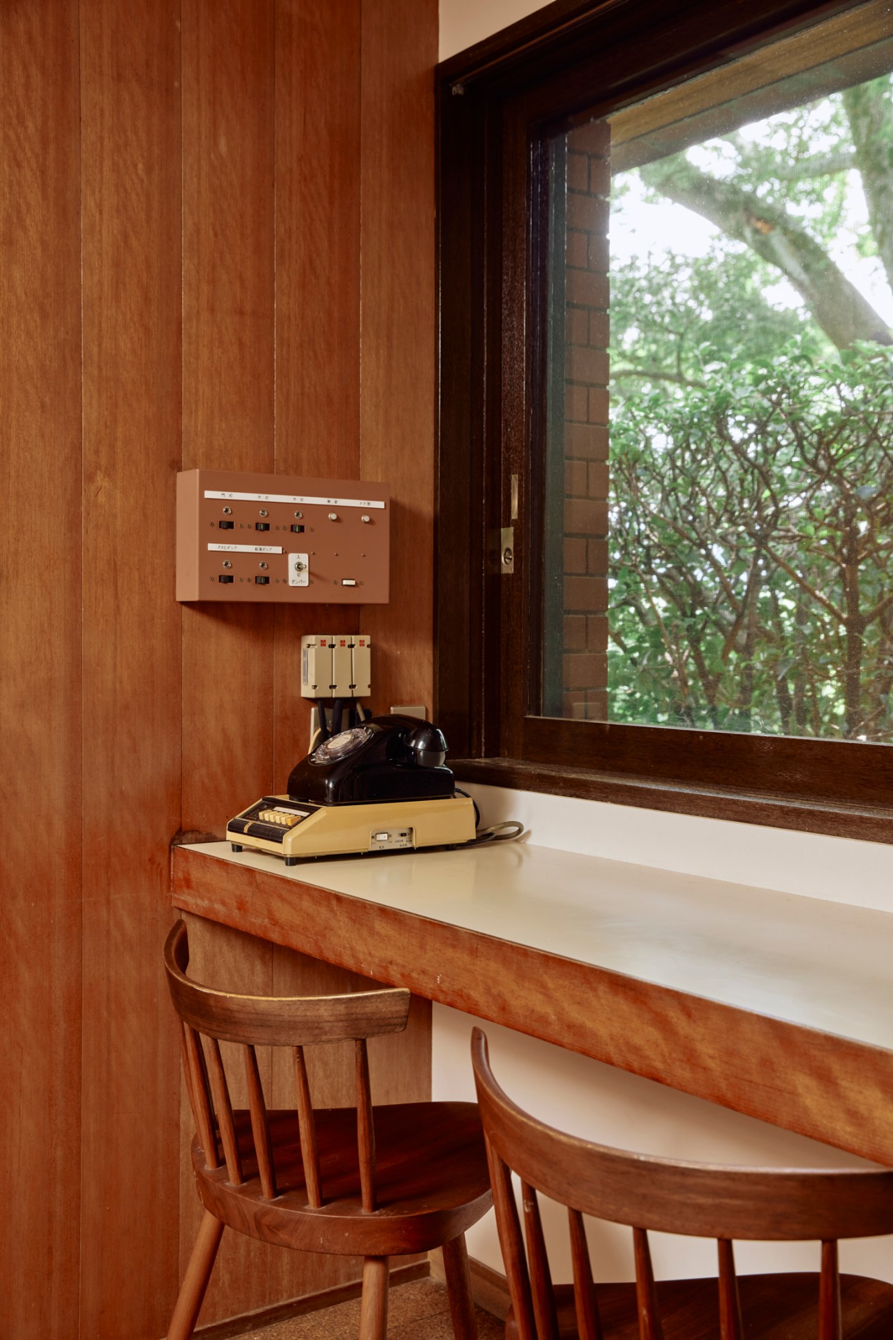 A vintage rotary telephone sitting on a wooden desk surface with control panels on the wall and a window view