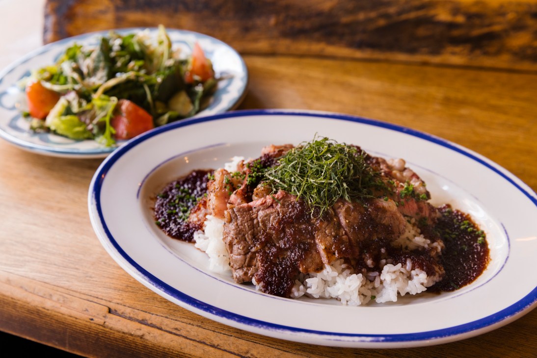Sliced grilled beef served over rice with dark sauce and green garnish alongside a fresh salad