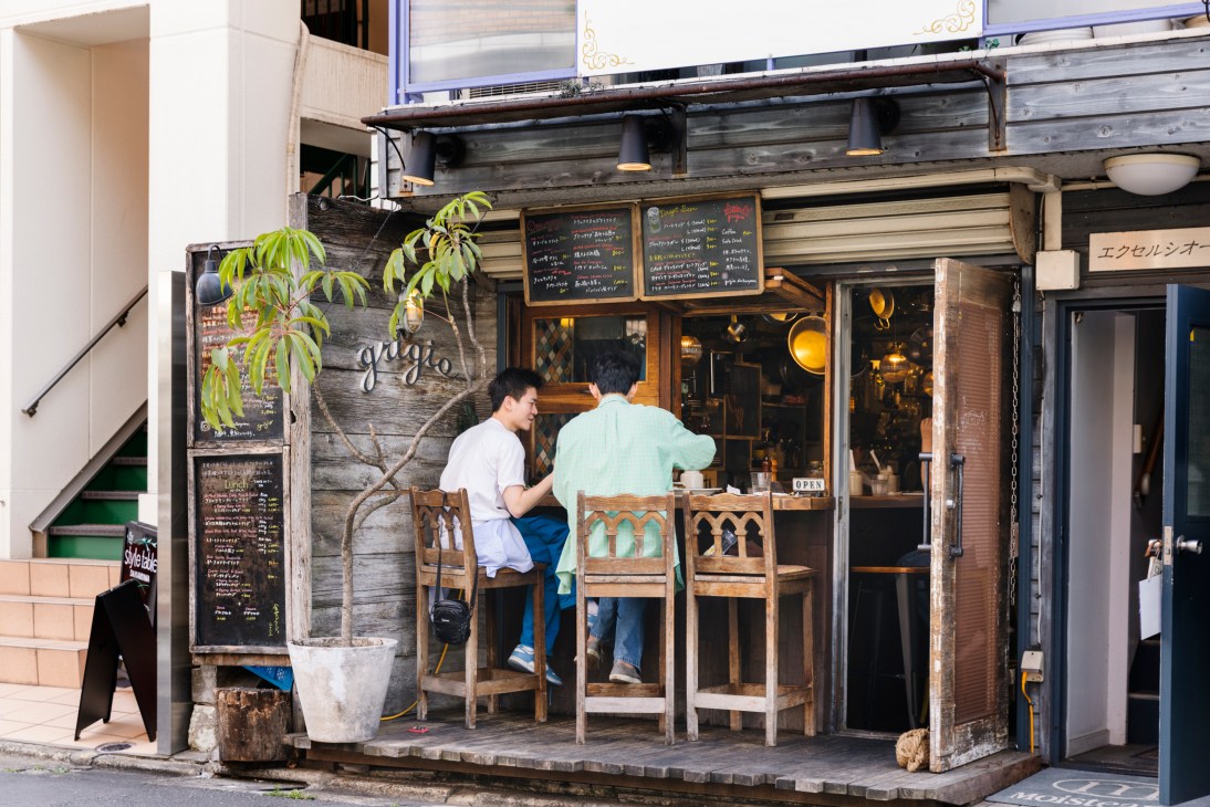Two men sit at a wooden counter outside a small restaurant with weathered wood exterior and menu boards