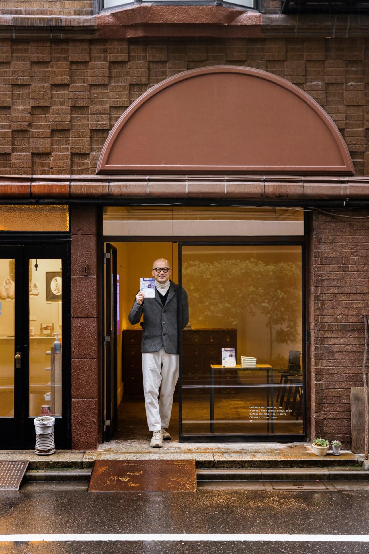 Man in glasses and grey cardigan holding book while standing in doorway of small Morioka Shoten bookshop