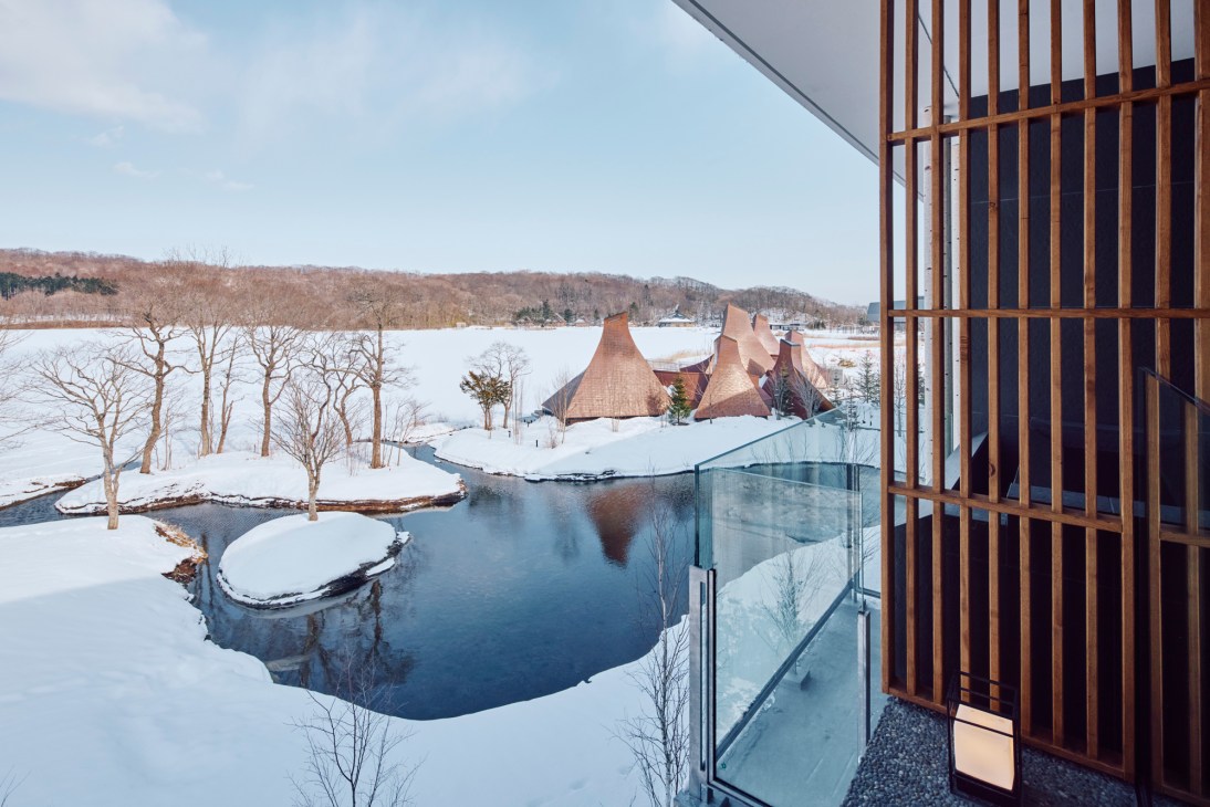 View from hotel balcony showing wooden screen, frozen Lake Poroto, and cone-shaped bathhouses in snowy winter landscape