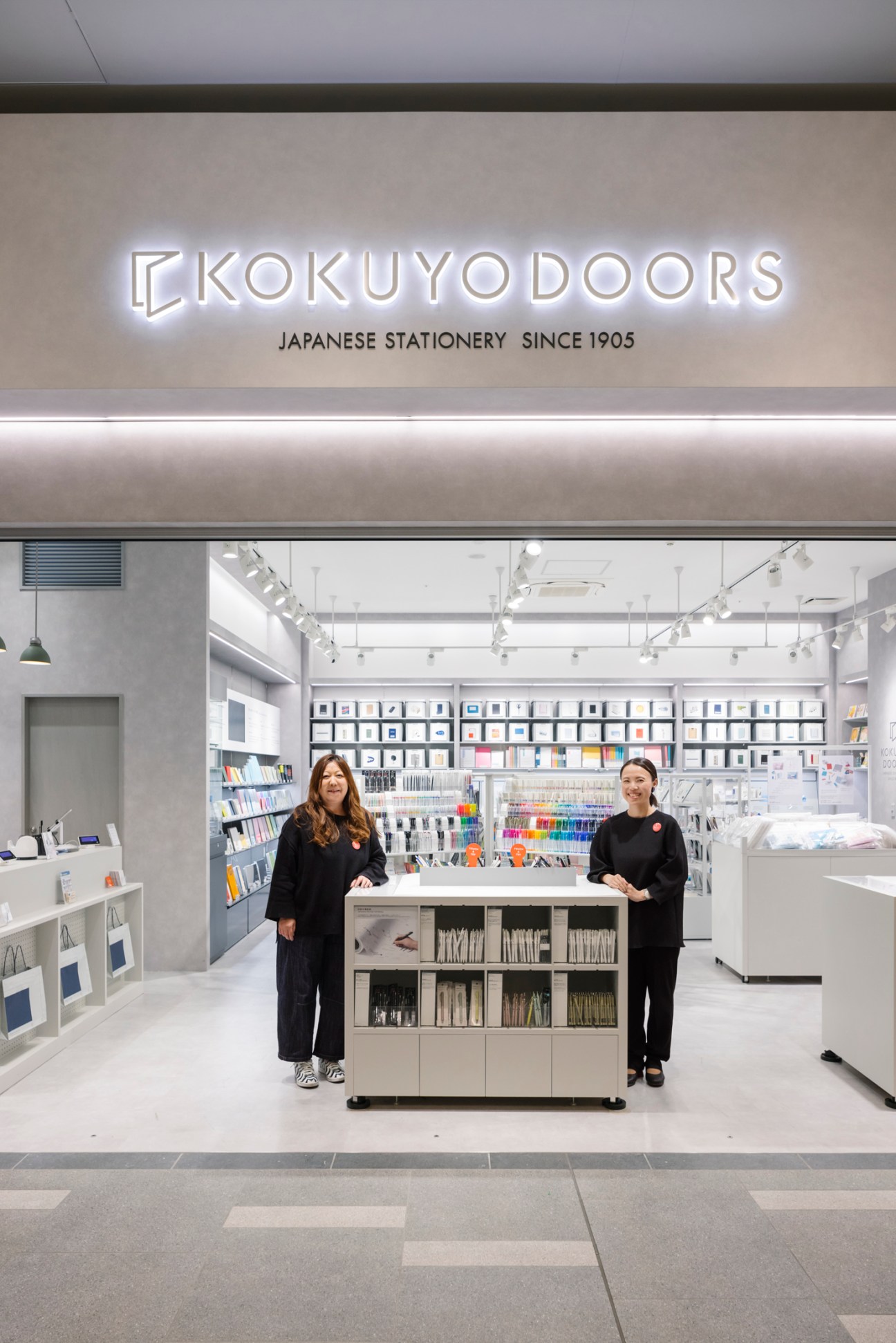Two staff members in black uniforms stand in a bright white Kokuyo Doors stationery store with organized displays