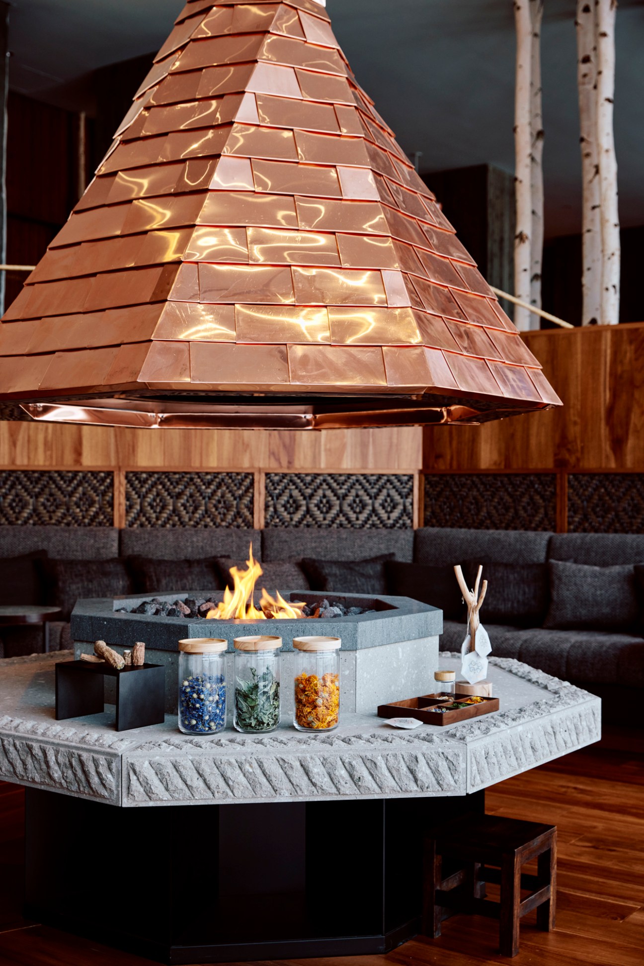 Modern hotel lobby with central stone fireplace featuring copper pyramid hood and surrounding seating areas