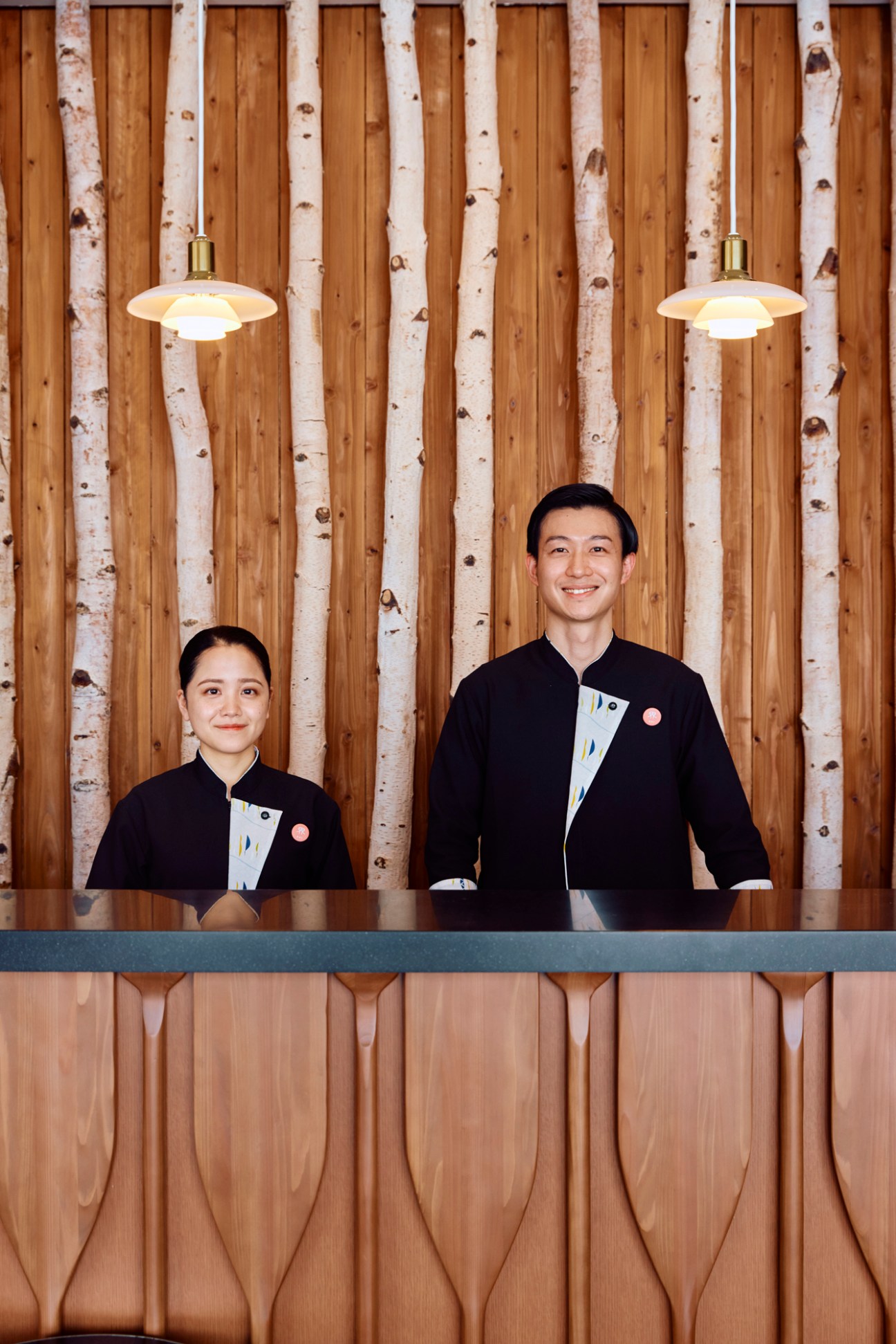 Two hotel staff members in black uniforms with mandarin collars standing behind wooden reception desk smiling