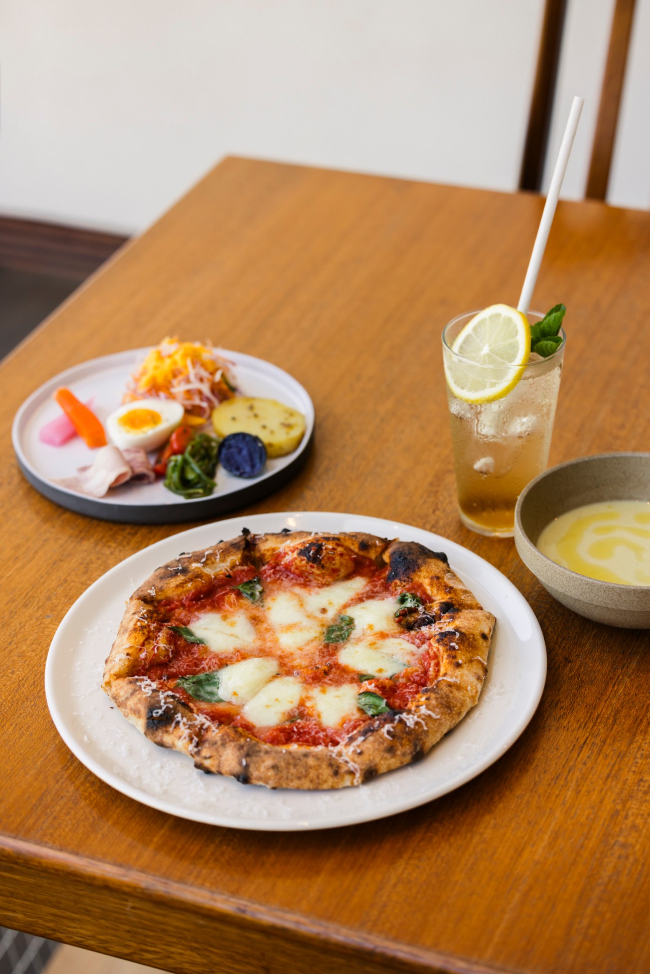 A Neapolitan pizza with fresh mozzarella and basil sits on a wooden table alongside appetizers and a lemon drink