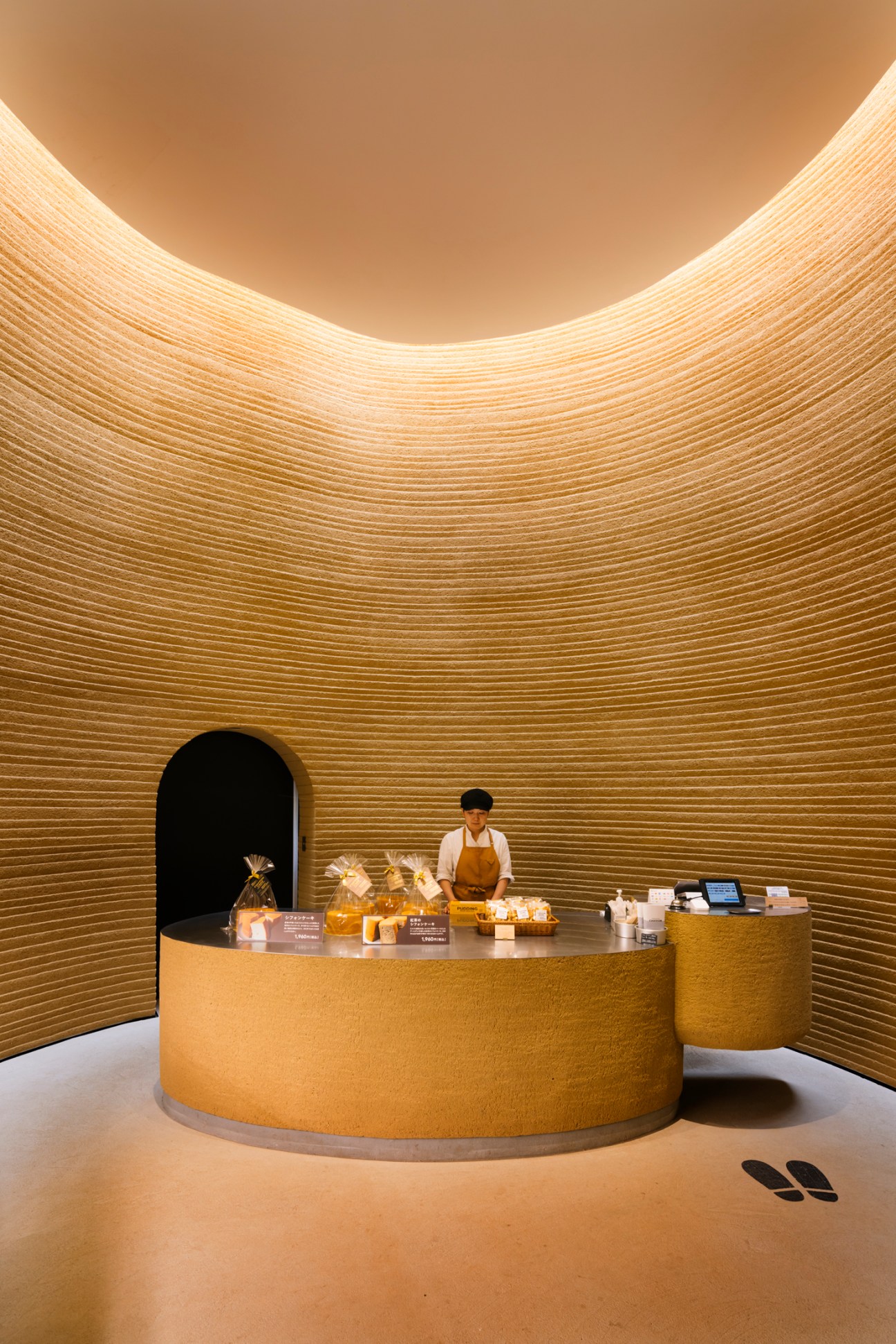 Staff member in apron stands behind curved counter in golden wood-lined bakery with arched ceiling and dark doorway