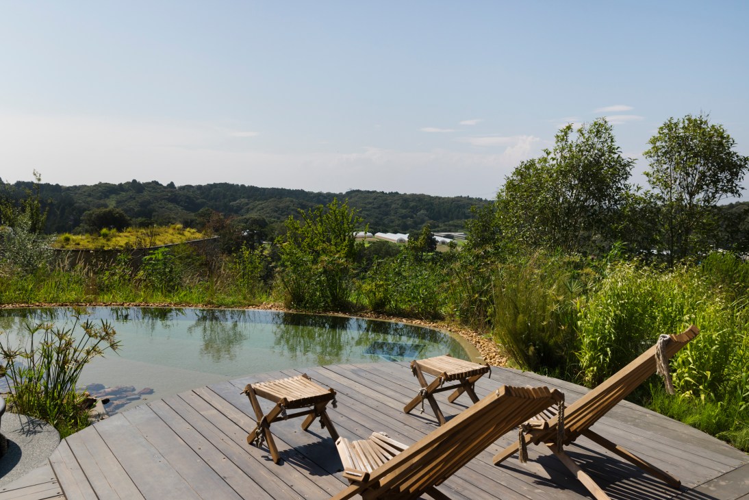 Wooden deck chairs and side tables beside a natural swimming pool overlooking forested hills in countryside