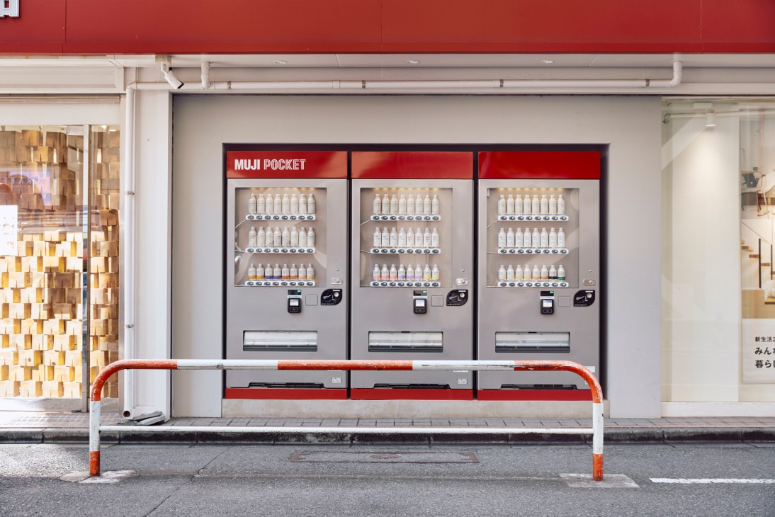 Three MUJI Pocket vending machines displaying small bottles on shelves, positioned in front of a storefront with a bench in the foreground.