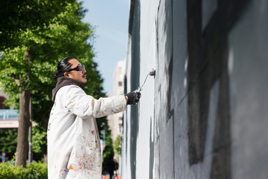 Person in white lab coat and sunglasses using paint roller on wall to cover graffiti