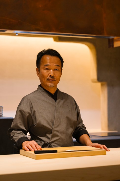 Chef Yoshinori Ishii stands behind a wooden cutting board in traditional Japanese chef attire at his restaurant kitchen