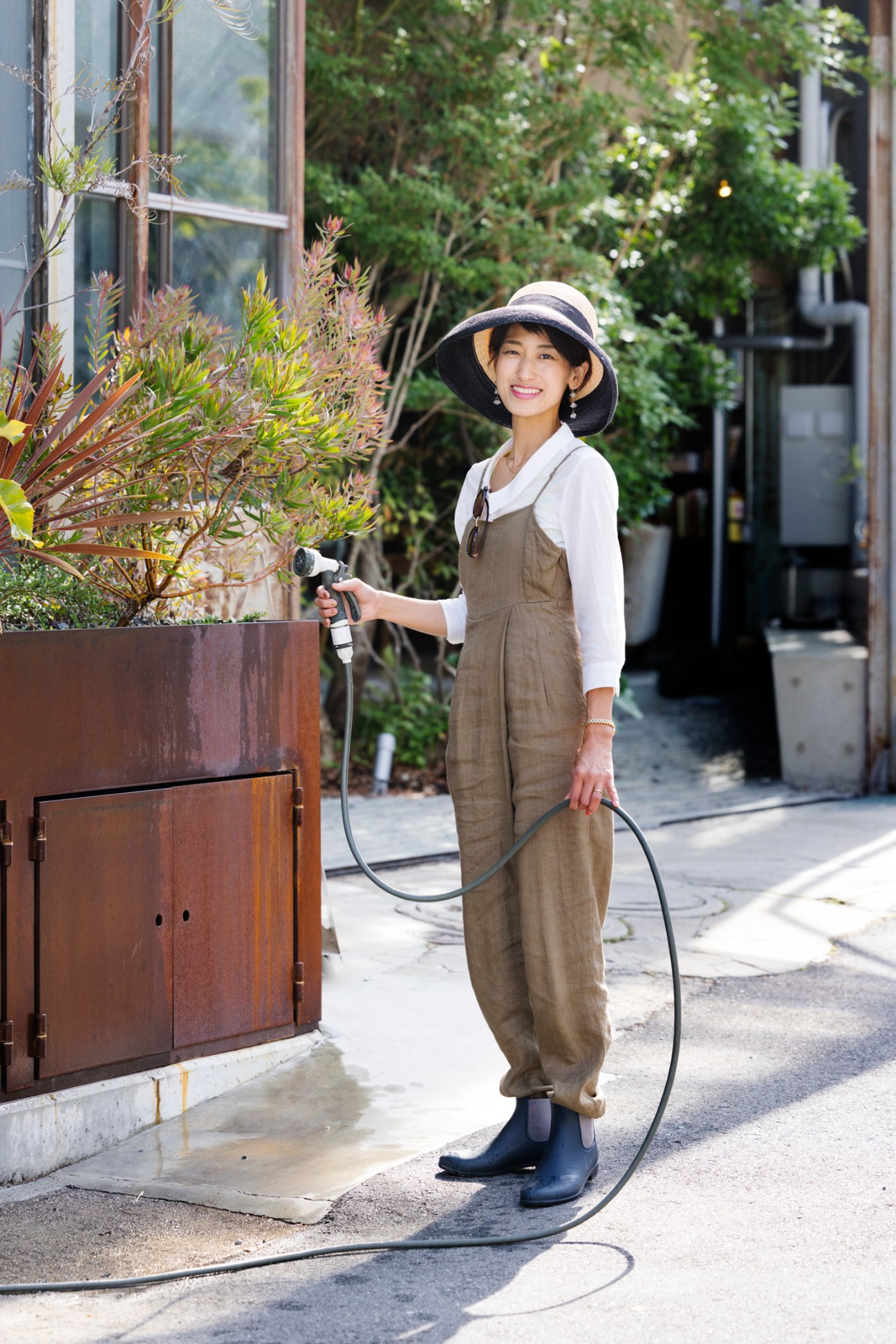Lan Haguma watering plants while wearing overalls and a wide-brimmed hat in outdoor garden area