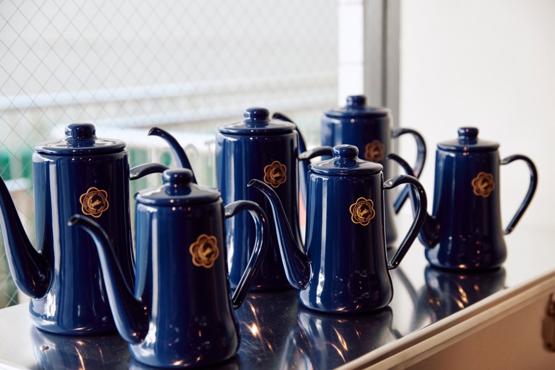 Six glossy navy blue enamel coffeepots with matching lids arranged on a reflective surface near a window