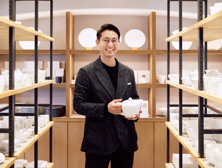 Naoki Mitani standing in porcelain showroom holding white bone china teapot surrounded by shelves of white tableware