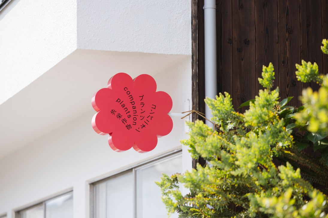 Red flower-shaped logo with Japanese text on white wall next to green foliage