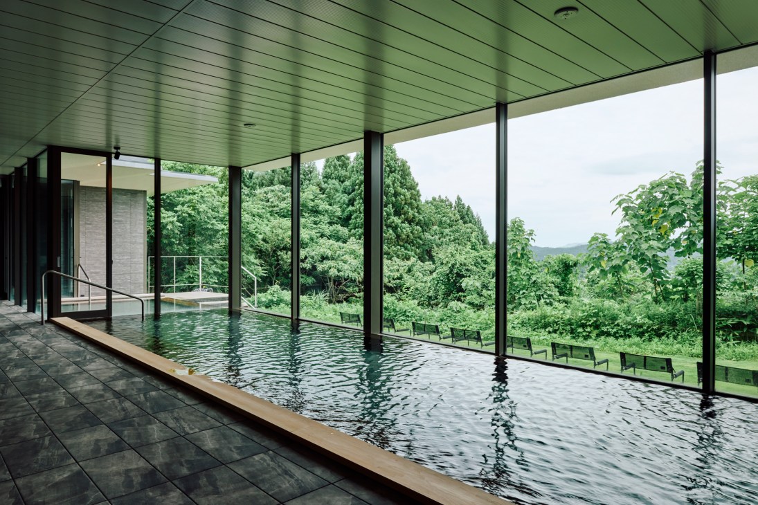 Men's hot spring bath with modern wooden interior, steaming water, and floor-to-ceiling windows overlooking lush green forest landscape