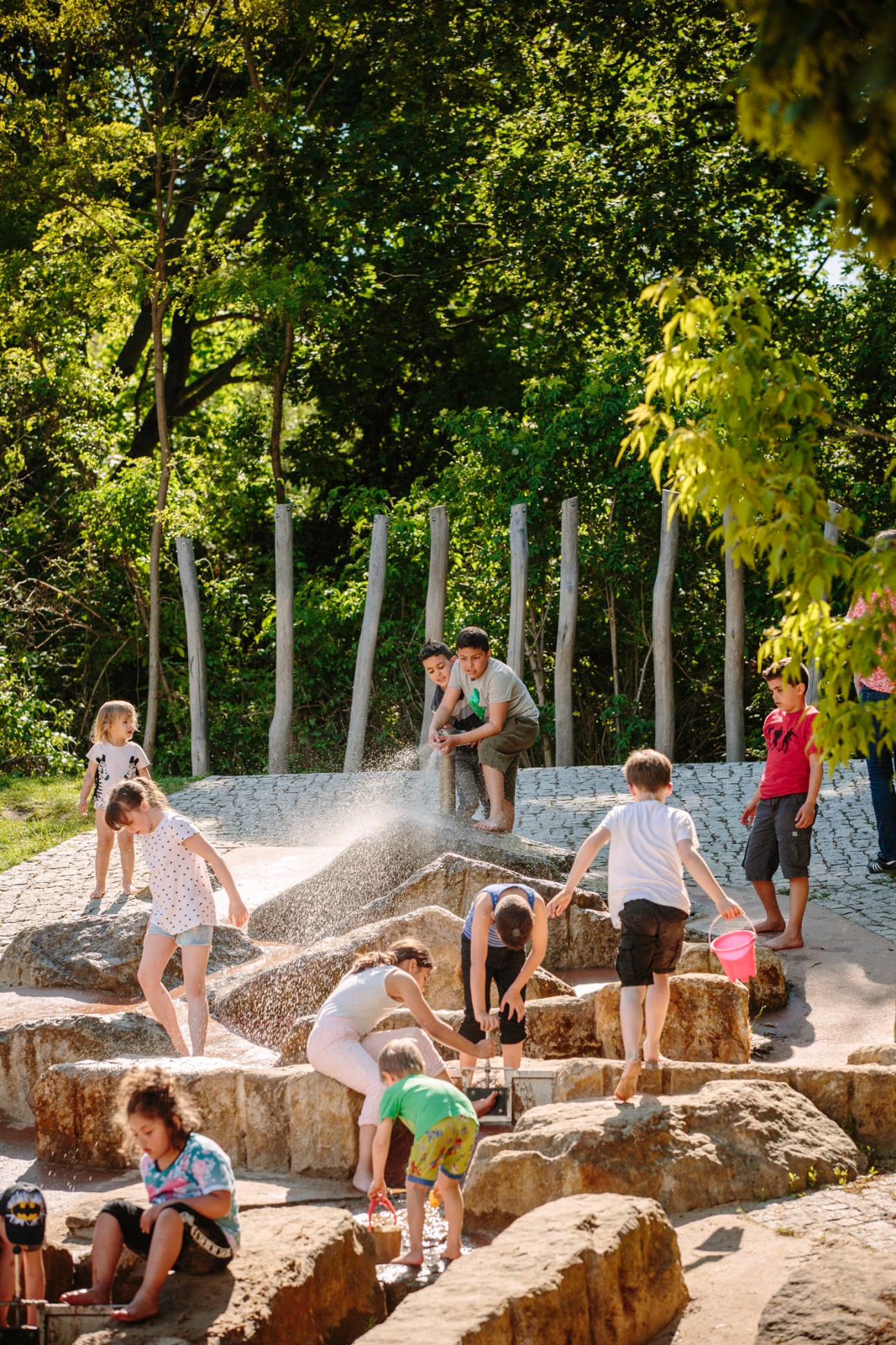 Children playing in a water feature at Park am Buschkrug in Neukölln, Berlin, with tall wooden posts and trees in the background.