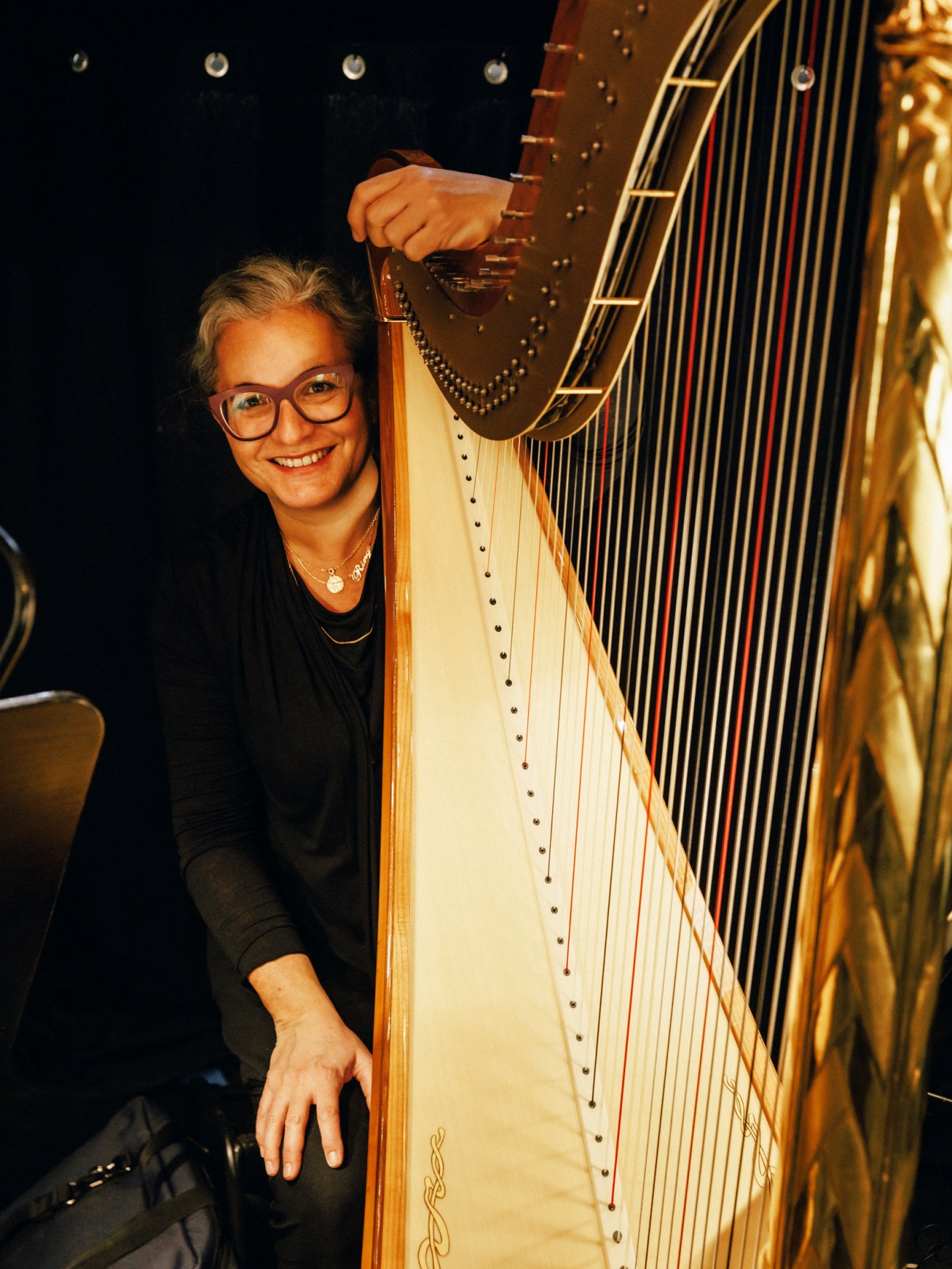 A smiling harpist wearing glasses poses beside a large golden harp on a dark stage.