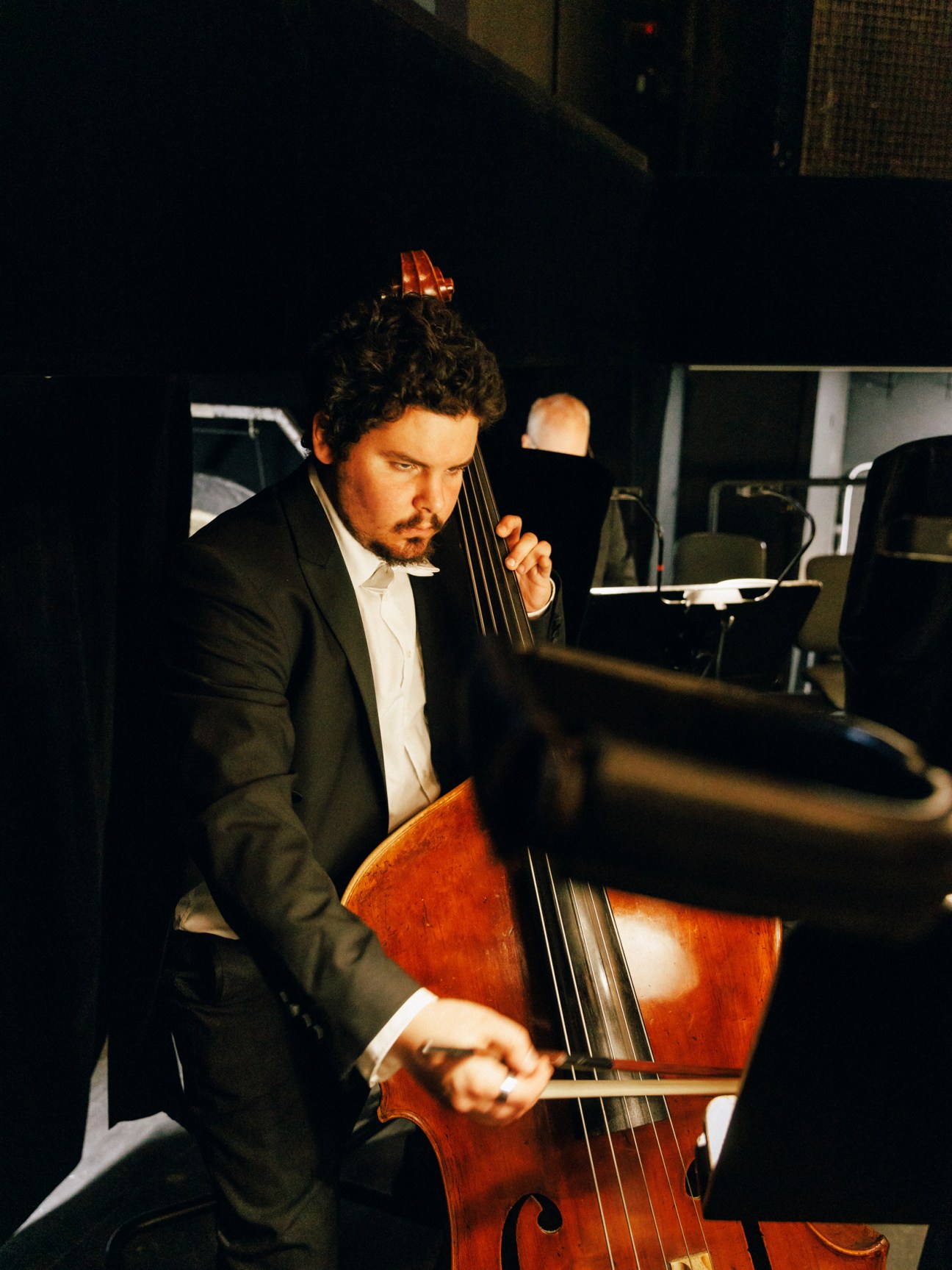 A cellist in formal attire focuses intently while playing in a darkened backstage area.