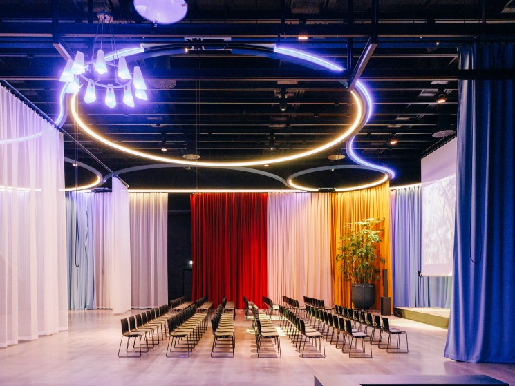 Modern event space with colorful curtain backdrop, curved neon lighting, and rows of black chairs arranged theater-style