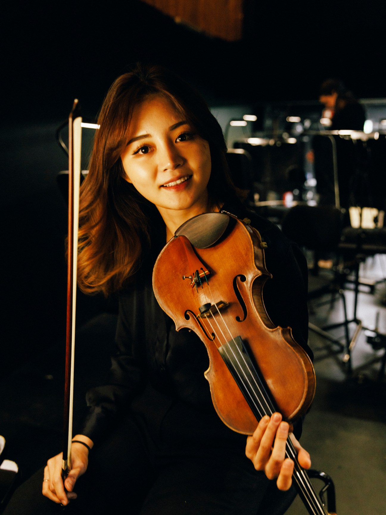 Female violinist holding a violin and bow, smiling at the camera in a backstage setting.