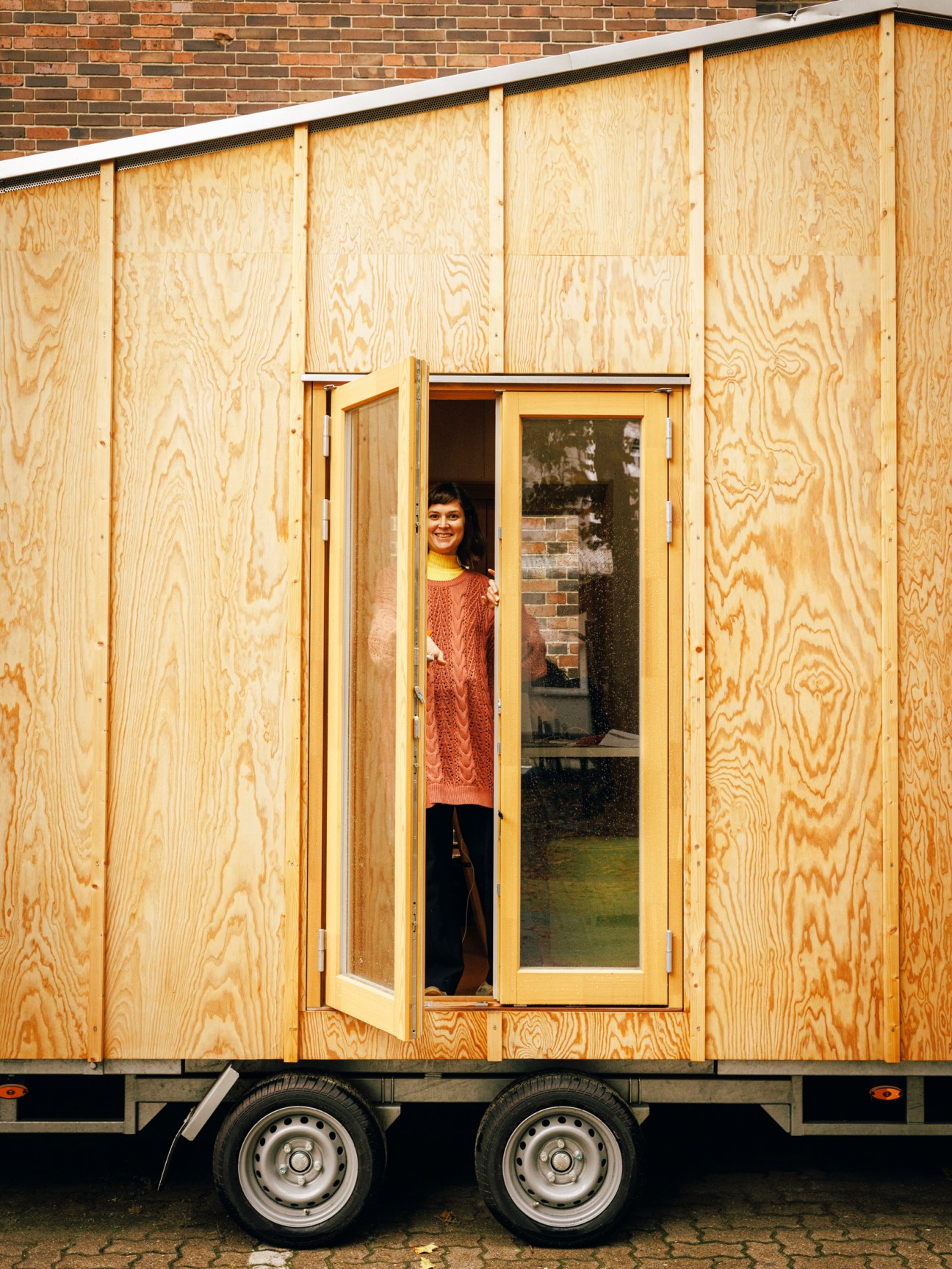 A woman in a rust-colored sweater stands in the doorway of a wooden mobile home or trailer with plywood siding
