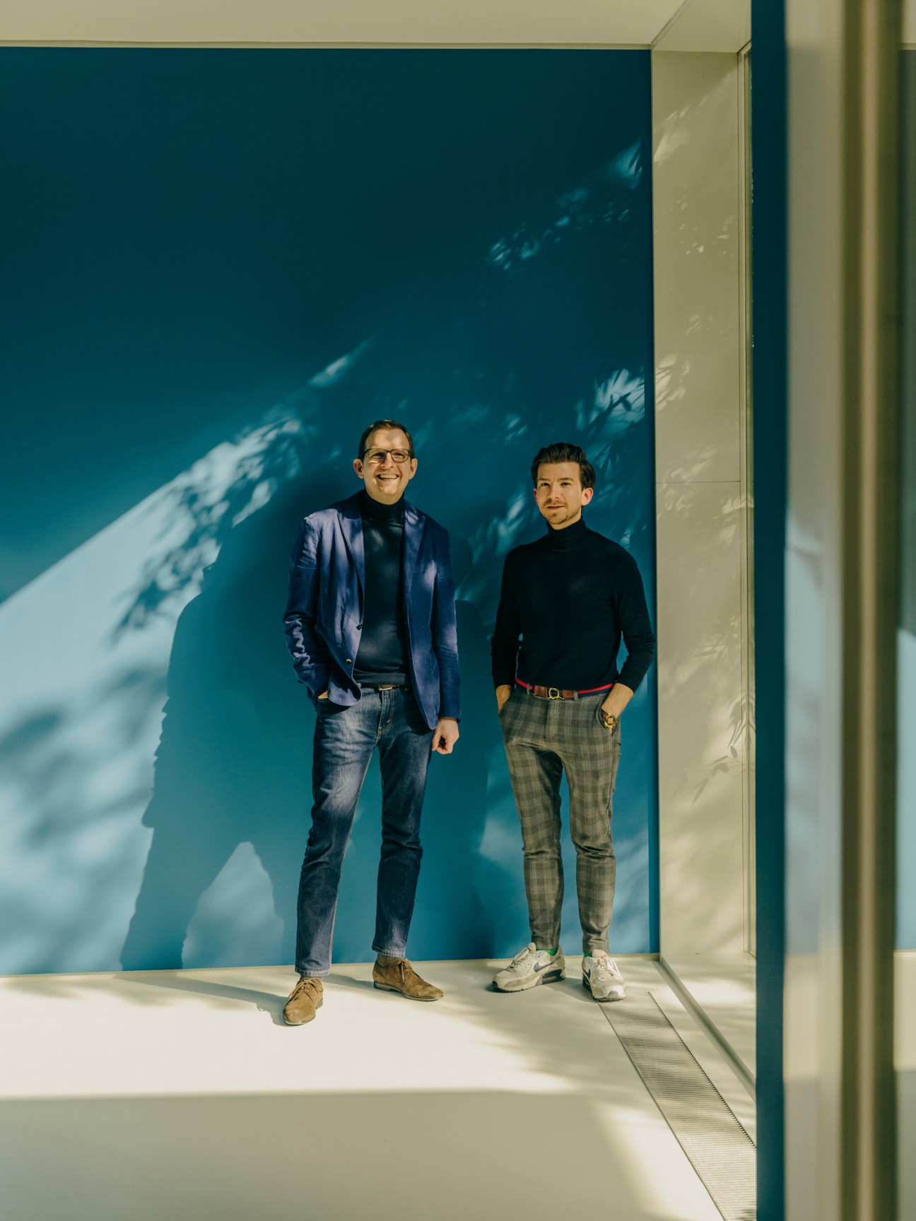 Two men stand in a minimalist gallery space with teal walls and natural light.