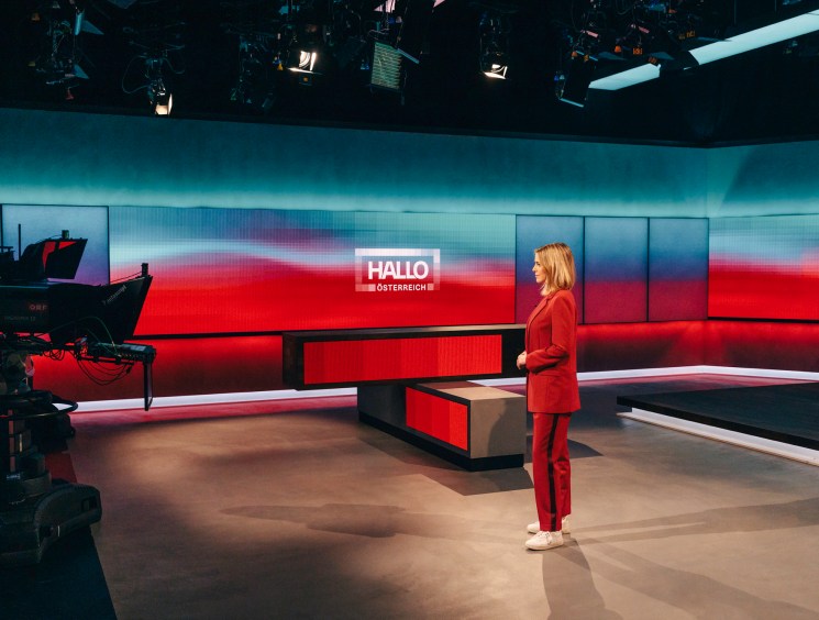 Television news anchor in red suit standing in modern studio with curved LED screens and professional lighting
