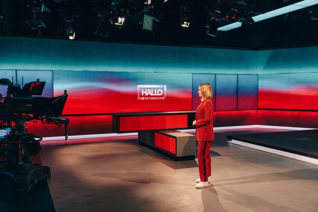 Television news anchor in red suit standing in modern studio with curved LED screens and professional lighting