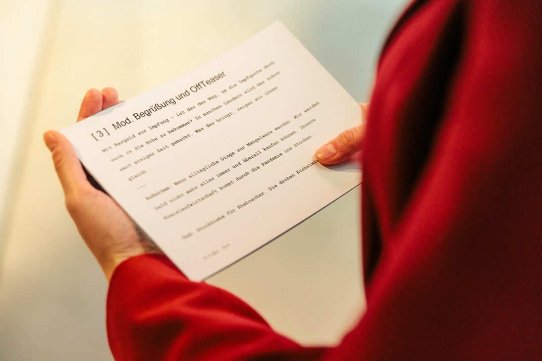 Person in red holding white paper with German text including 'Mod. Begrüßung und Off Teaser'