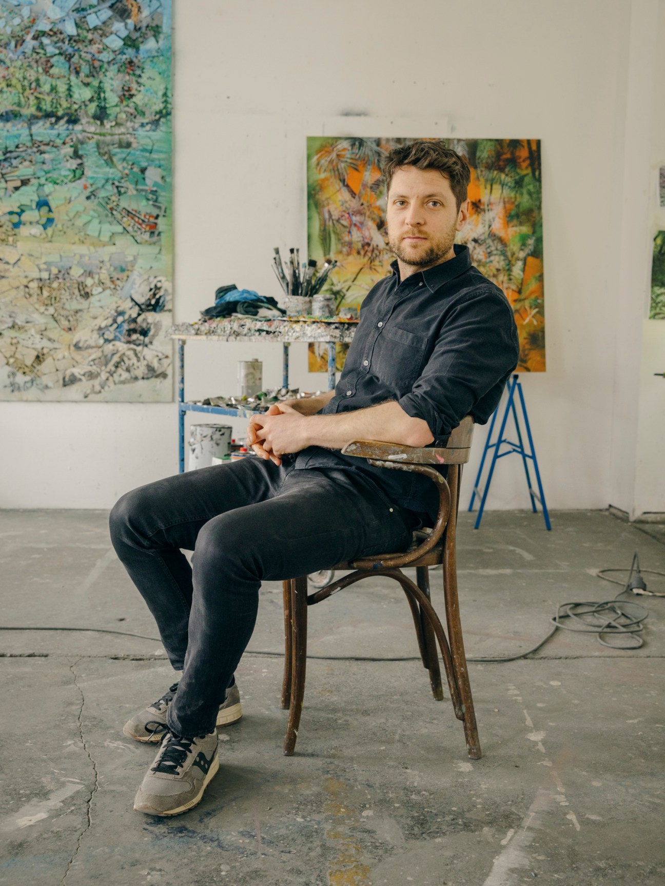 Artist Philipp Fürhofer sits in a studio chair holding a palette, surrounded by colorful abstract paintings.