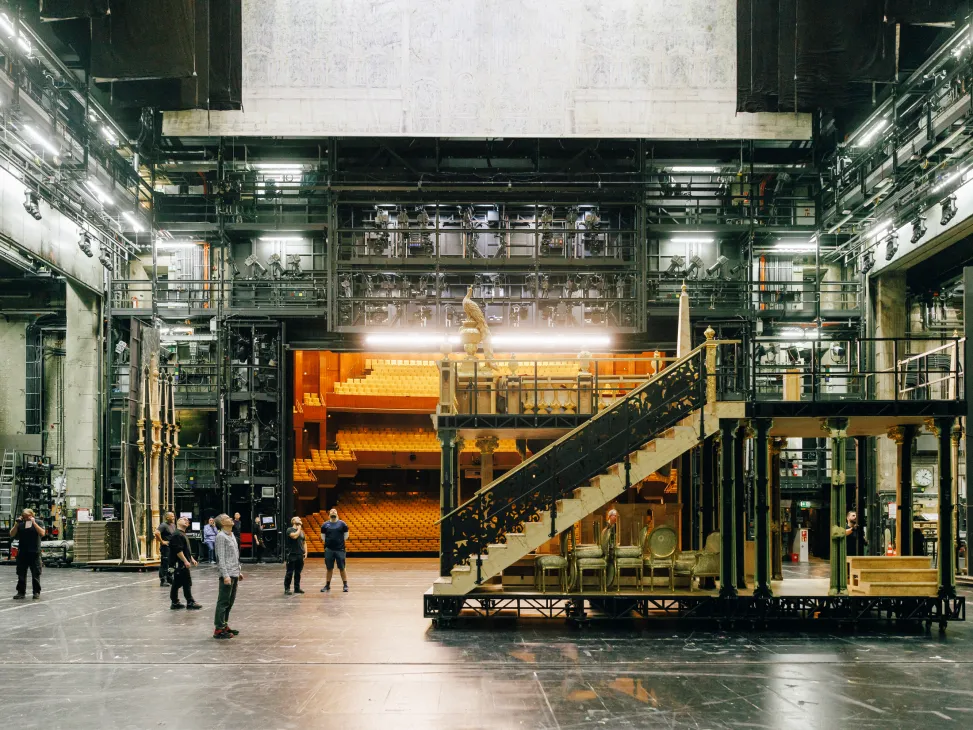 Backstage area of Deutsche Oper with industrial scaffolding, metal catwalk stairs, and orange auditorium seating visible in background.