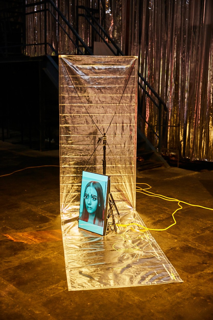 Contemporary art installation with clear plastic sheeting, projected portrait, and yellow cables in industrial gallery space