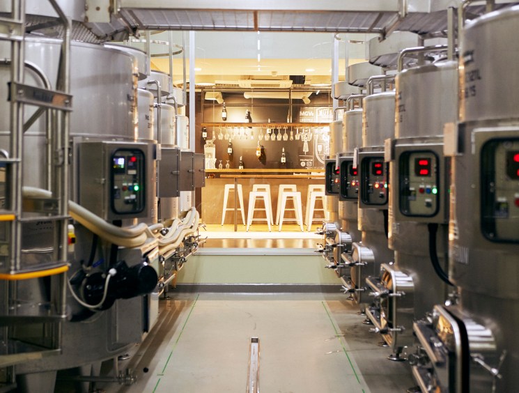 A narrow corridor between stainless steel brewing tanks leading to a warmly lit craft beer bar with bar stools.