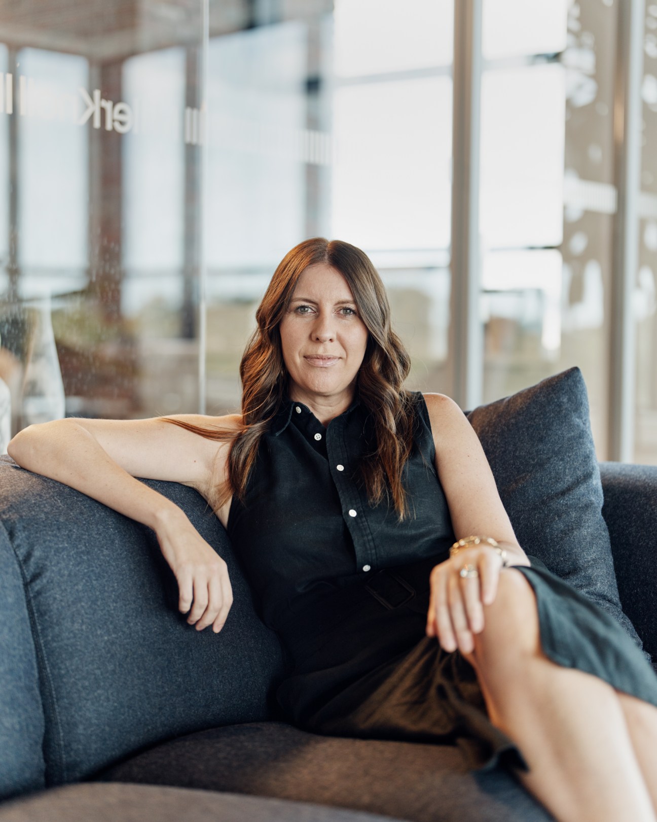 Woman with long brown hair wearing black sleeveless shirt sitting on gray couch in bright office space