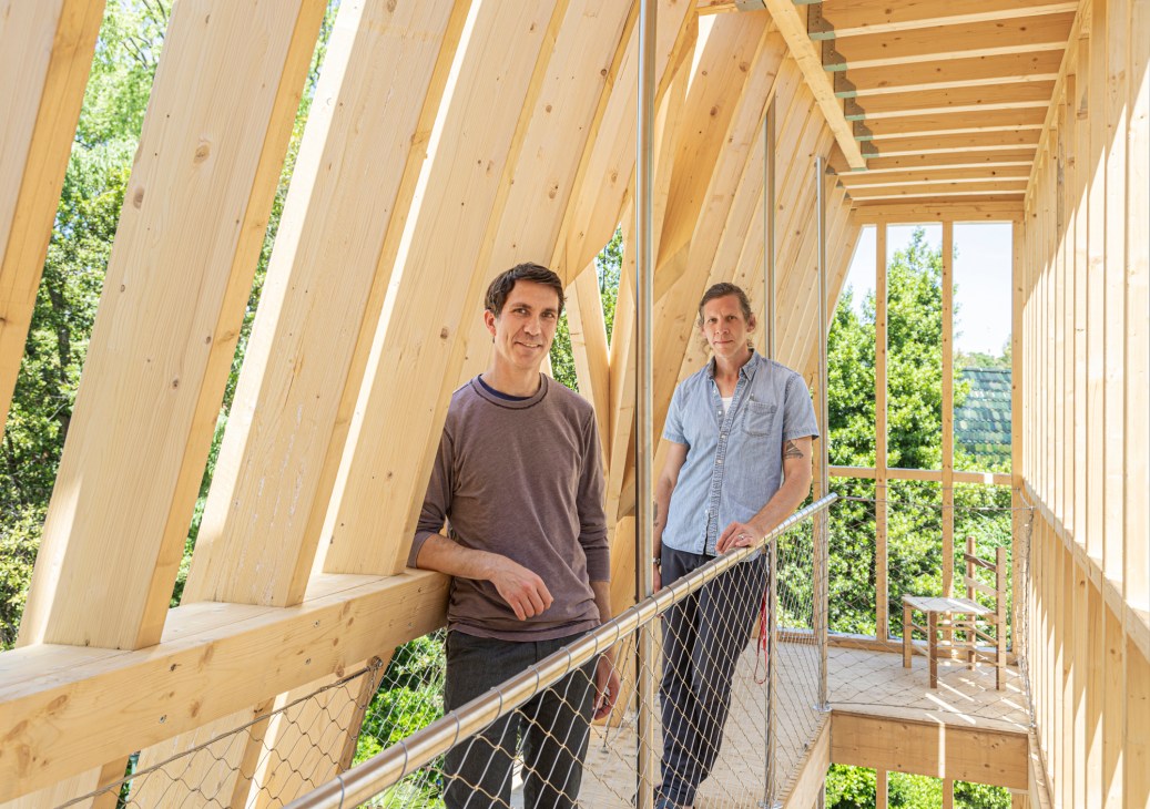 Two men stand in a modern wooden structure with angled timber beams and metal railings, surrounded by green vegetation.
