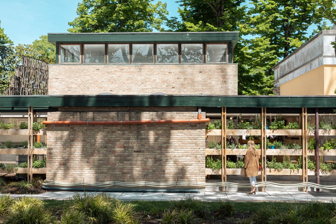 Modern brick building with green roof and wooden plant shelving structure in a garden setting.