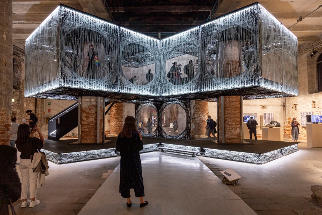 Illuminated installation with string-like fibrous structure featuring eye motifs displayed in a brick industrial space at Venice Architecture Biennale.