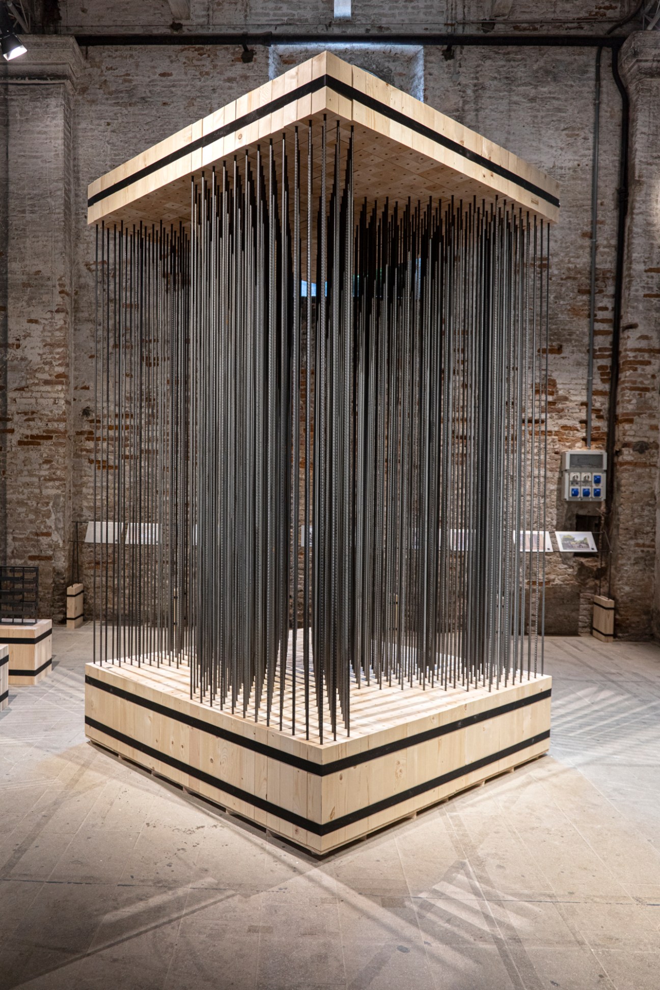 Wooden and metal latticed pavilion structure with peaked roof in industrial gallery space.