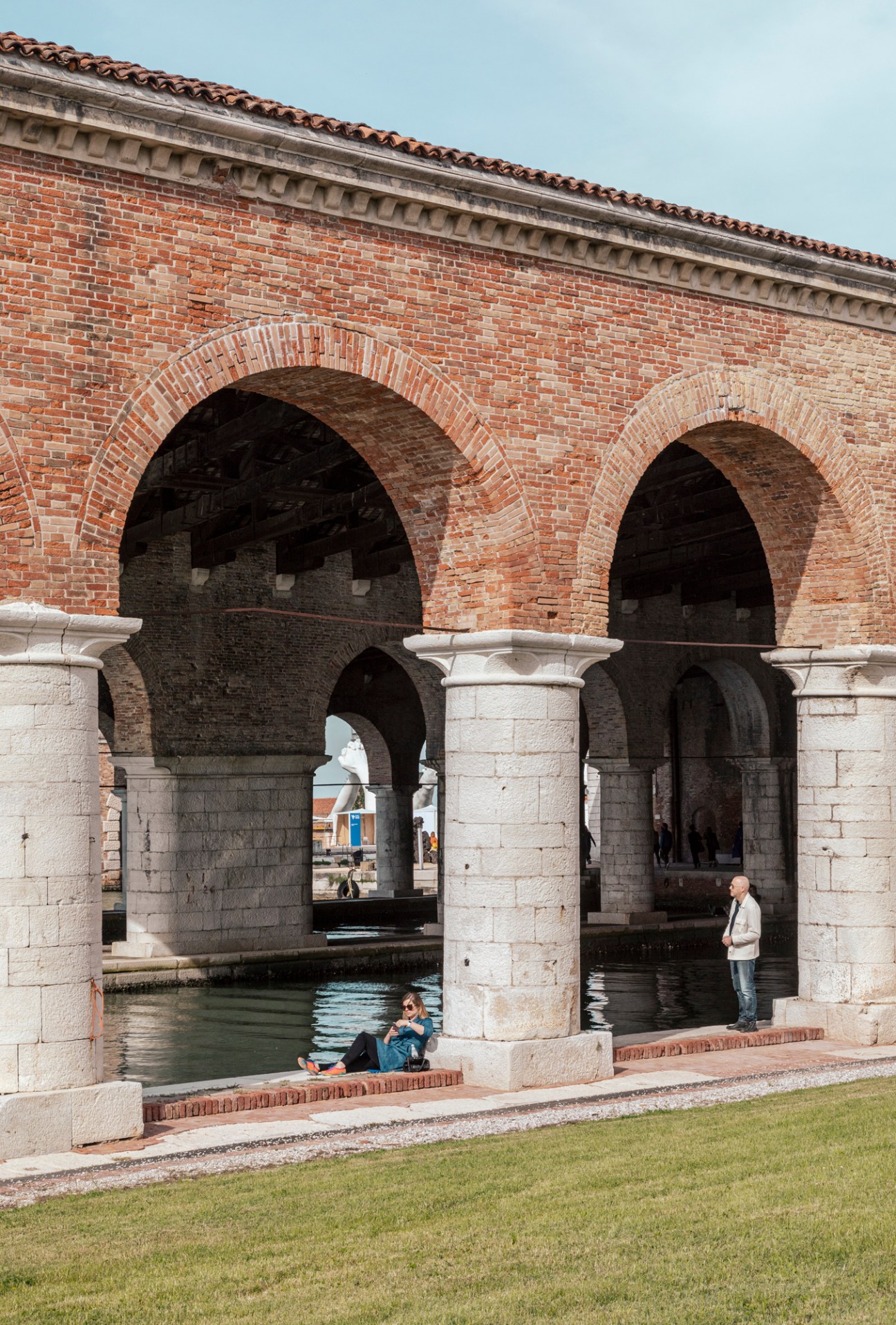 Architectural arches with columns frame a waterway beneath a brick building with ornate cornice detail.