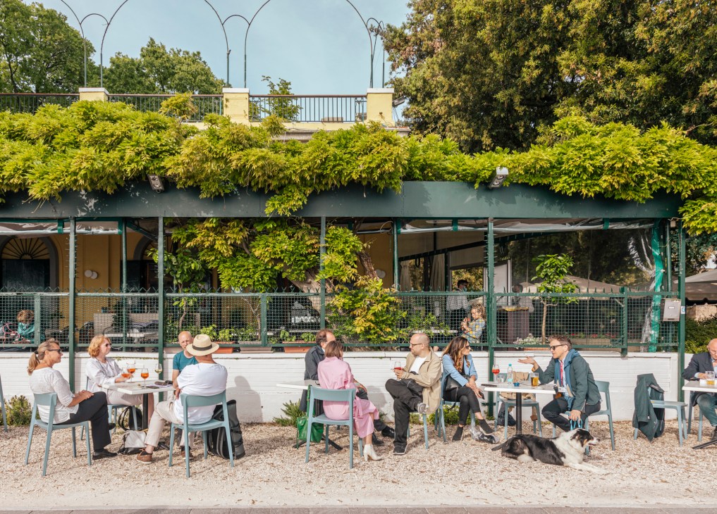 People seated at outdoor tables in front of a green-roofed café structure with climbing ivy and arched street lamps overhead.