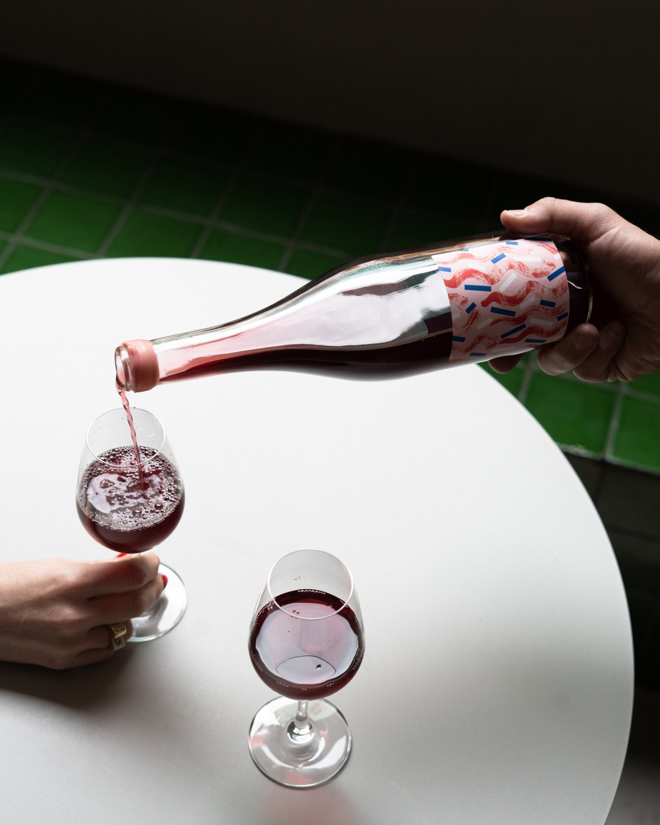 Hand pouring red wine from a decorative bottle into a glass on a white table with another glass nearby