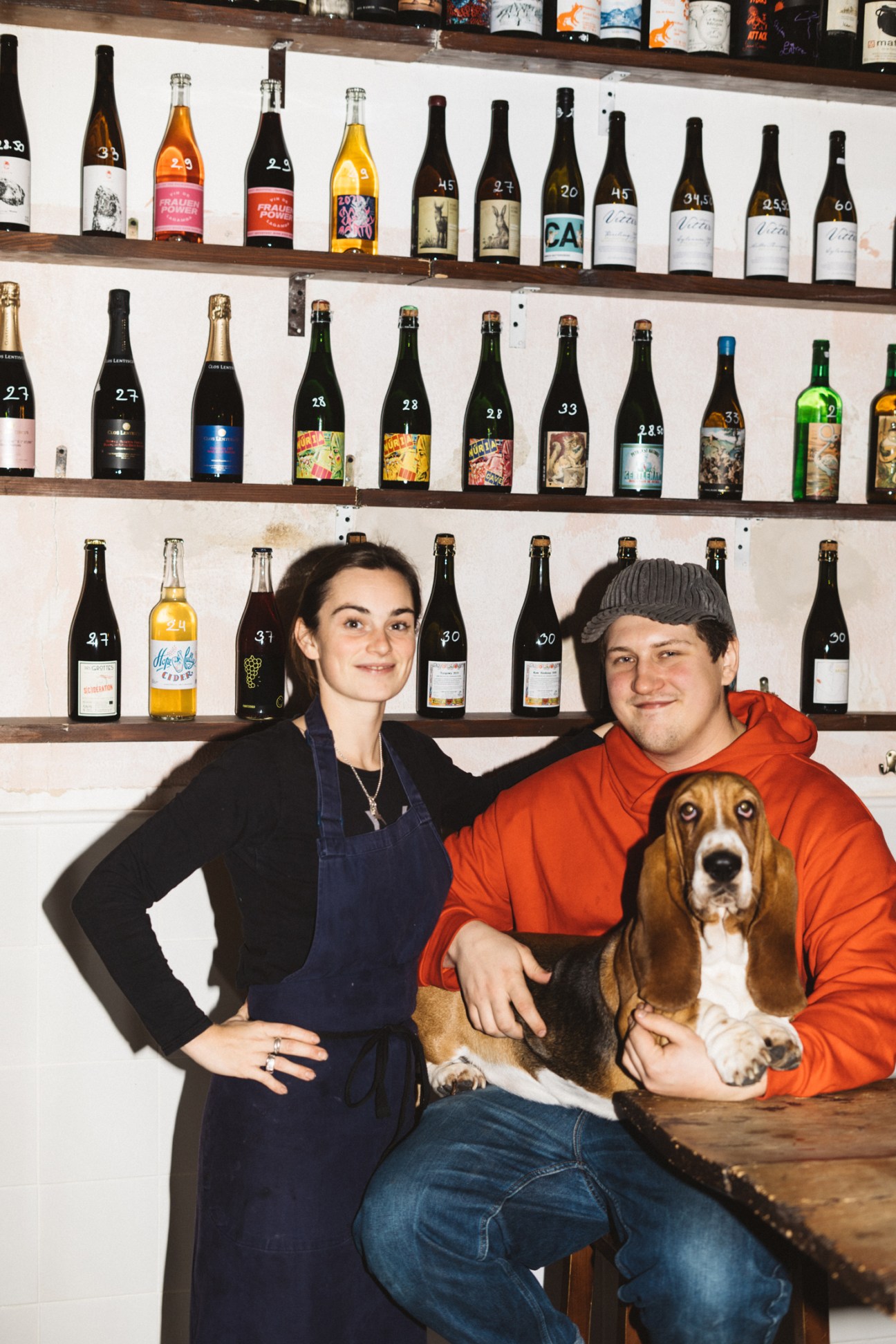 Woman and man sitting in front of shelves of wine bottles with a basset hound dog