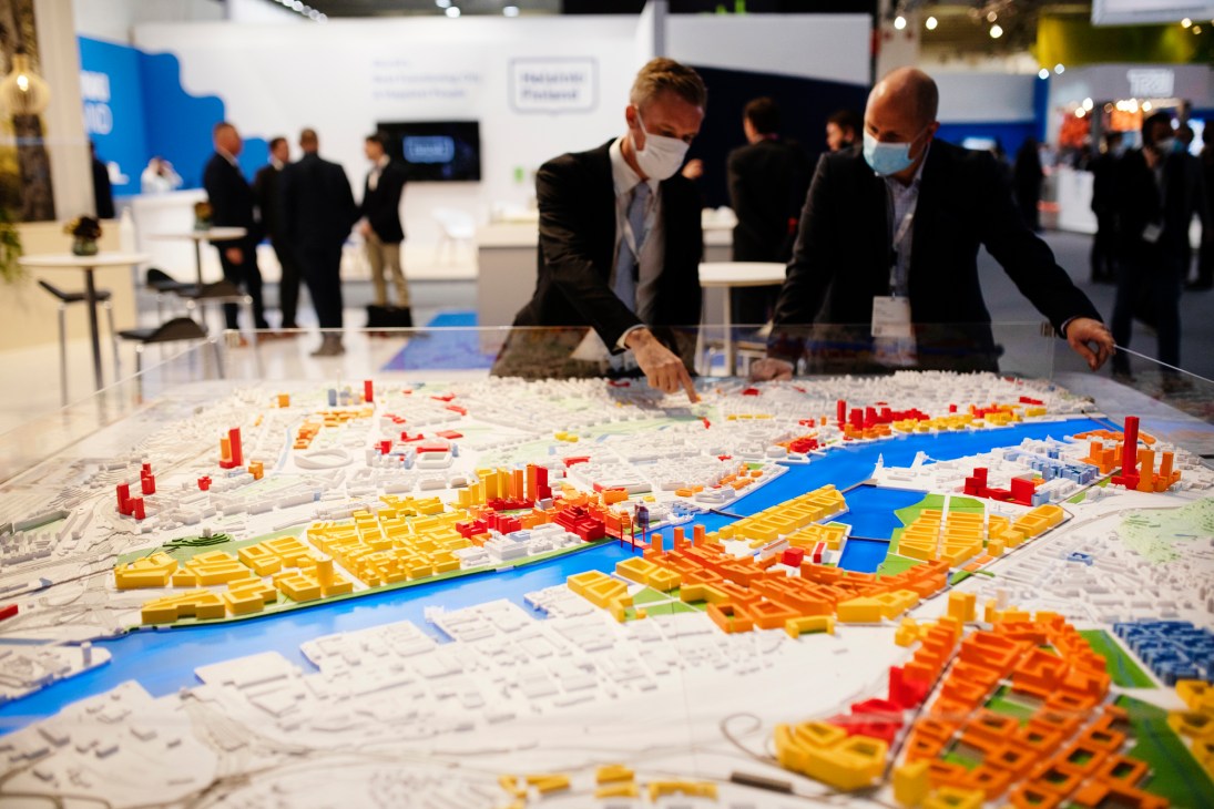 Two businessmen in masks examining a detailed architectural model with colorful building blocks at a trade fair