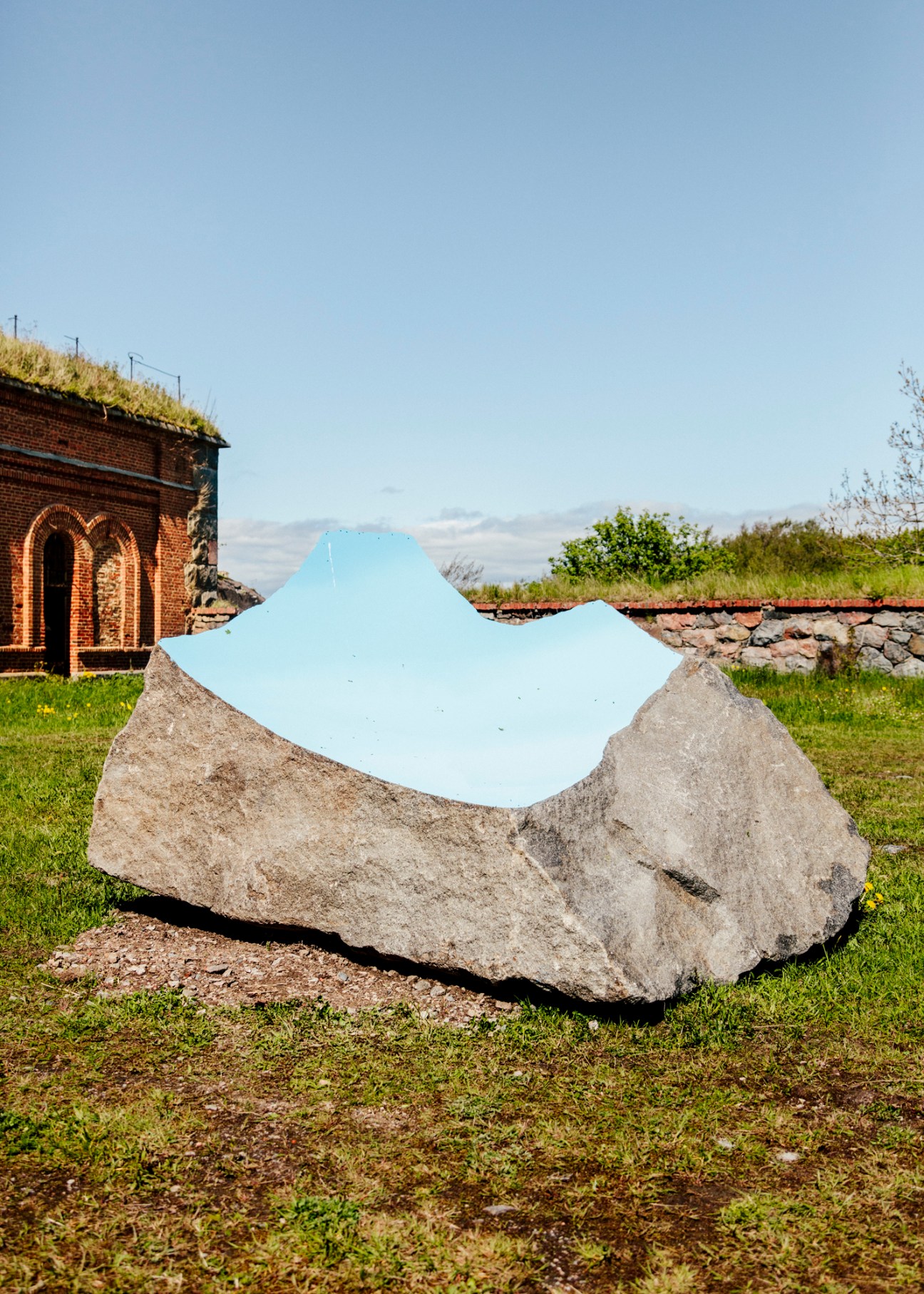 A large light blue mirror sculpture sits on a grey boulder in a grassy field with an old brick military building behind it.