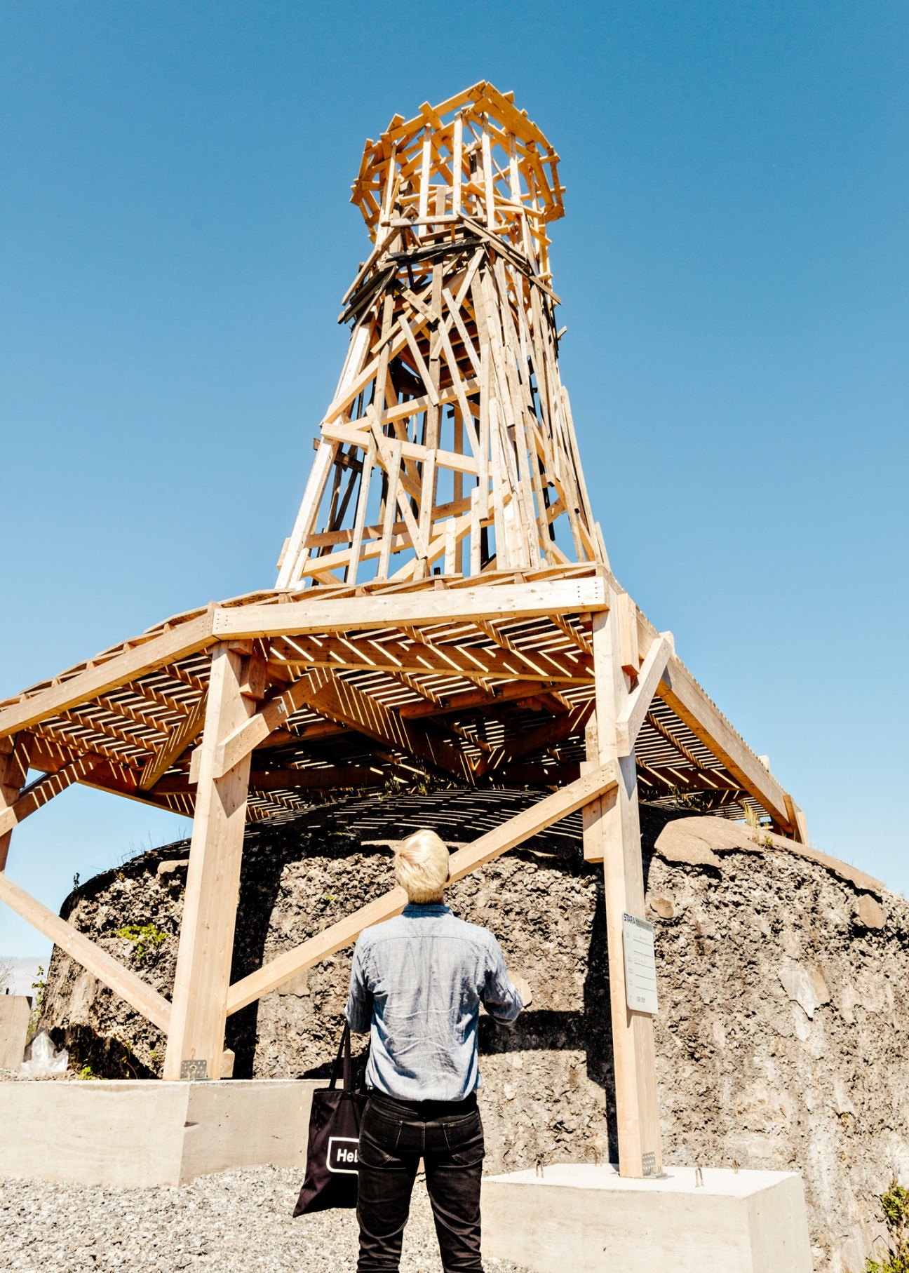 A visitor stands before a tall wooden lattice tower structure with geometric patterns, built on a pile of gravel against a clear blue sky.