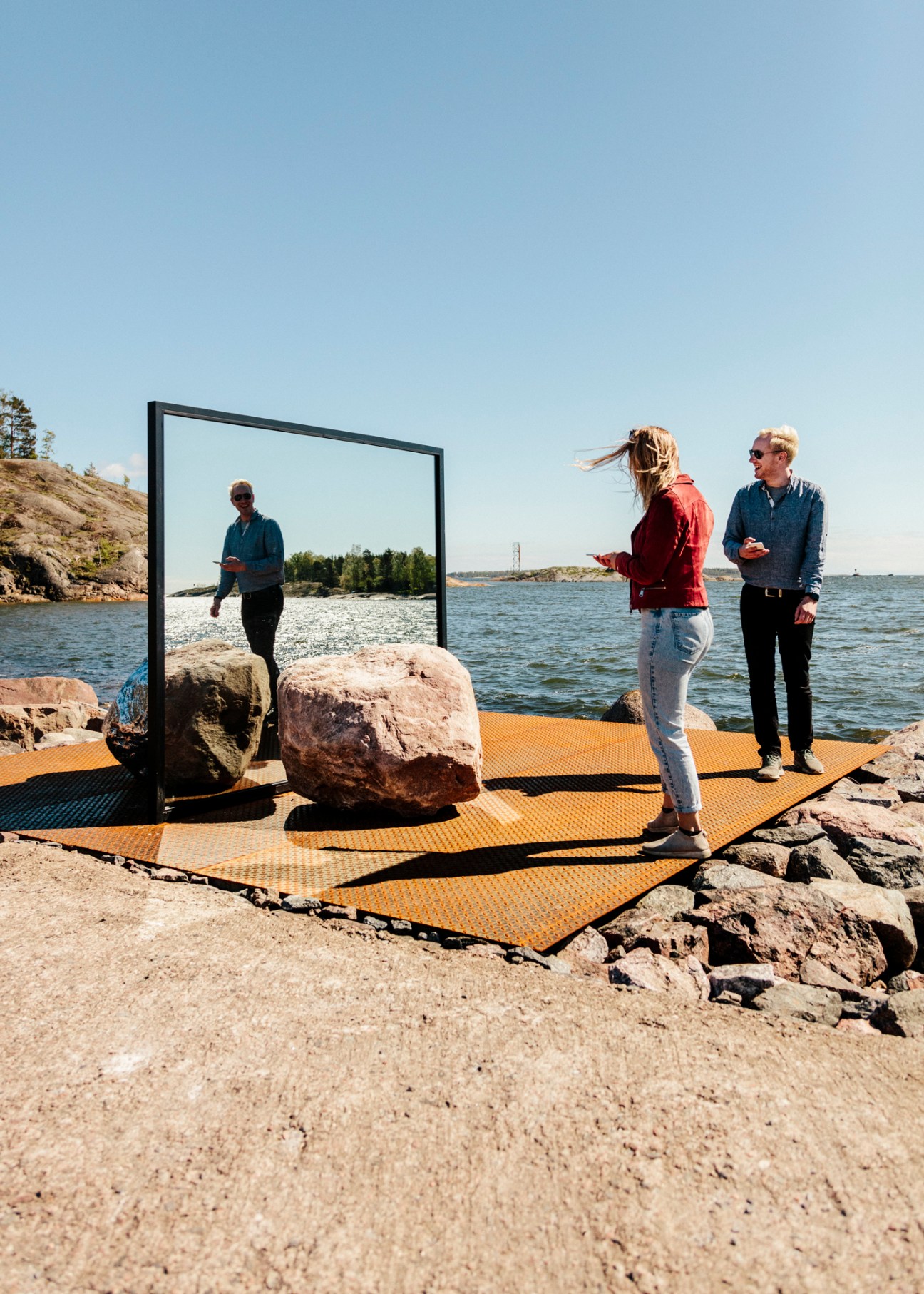 Visitors viewing Alicja Kwade's 'Big Be-Hide' sculpture featuring a black frame mirror and boulder on a rocky peninsula by the sea.