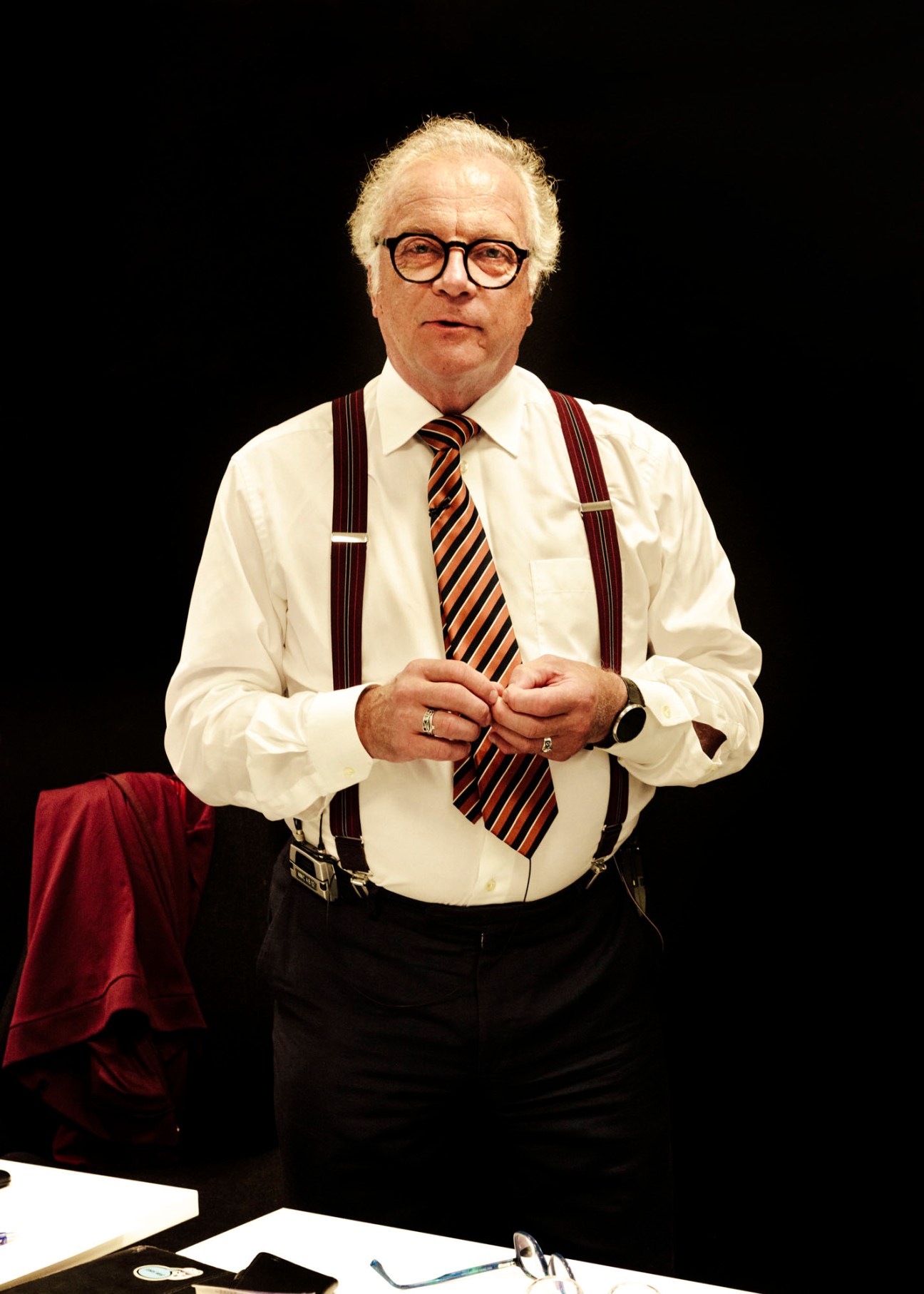 Matti Rönkä standing in formal attire with suspenders and striped tie against dark background