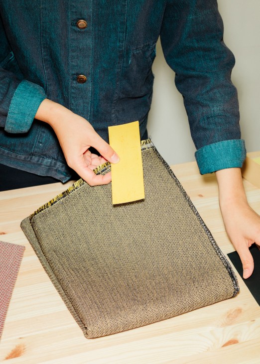 Person wearing denim jacket holding yellow card above textured fabric sample on wooden surface.