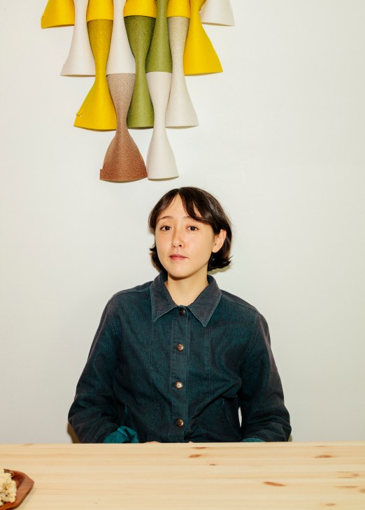 Elisa Defossez sits at a wooden table beneath conical textile designs in yellow, green, cream and brown tones.