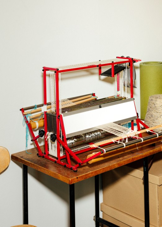 Red loom with white thread and wooden weaving implements on wooden table.